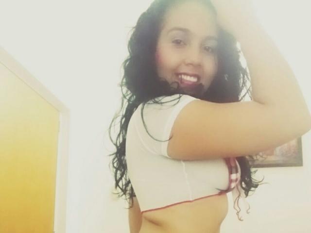 JadeTylerHot with long black wavy hair, smiling, wearing a white crop top, exposing her tits, raising her arm, standing in a brightly lit room, looking over her shoulder.
