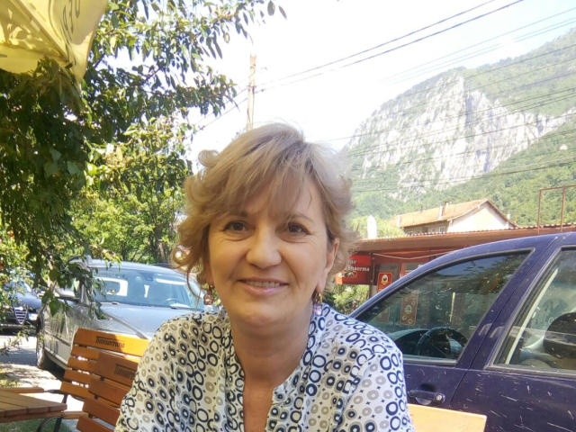 ExperiencedAlana with short, curly blonde hair, smiling, wearing a white patterned shirt, sitting outside, in front of a purple car, on a sunny day, with mountains in the background.