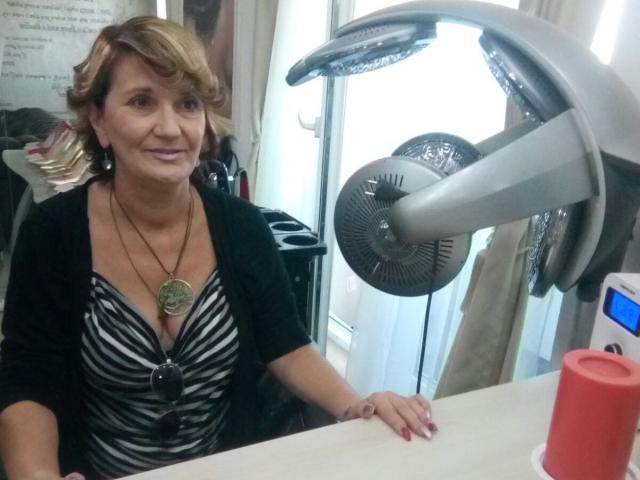 ExperiencedAlana, middle-aged woman with short blonde hair, wearing a black cardigan over a zebra-striped top, sitting next to a large hairdryer, red cup on the counter.