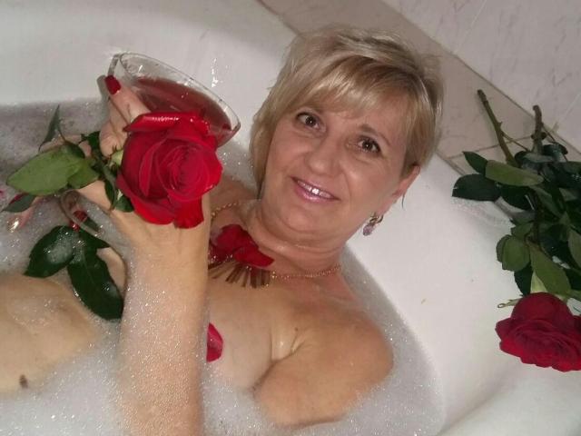Alana, experimentada, en una bañera llena de burbujas, sosteniendo una rosa roja en una mano y una copa de vino tinto en la otra, sonriendo, con cabello rubio corto..