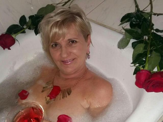 ExperiencedAlana in a bathtub with rose petals, holding a wine glass, wearing a gold necklace, smiling, with roses in the water and leaves around her.