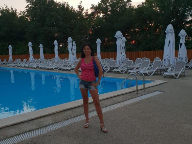 MadameAlexaX se tient près d'une piscine, vêtue d'un haut rose, de shorts en jean et de sandales, avec un sac à dos bleu, montrant ses fesses et ses seins, dans un cadre extérieur et décontracté..