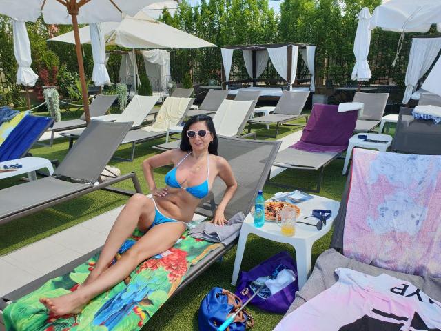 MadameAlexaX con un bikini azul, acostada en una tumbona con las manos sobre la barriga, gafas de sol puestas y una toalla de colores debajo. Tumbonas, sombrillas y bebidas alrededor..