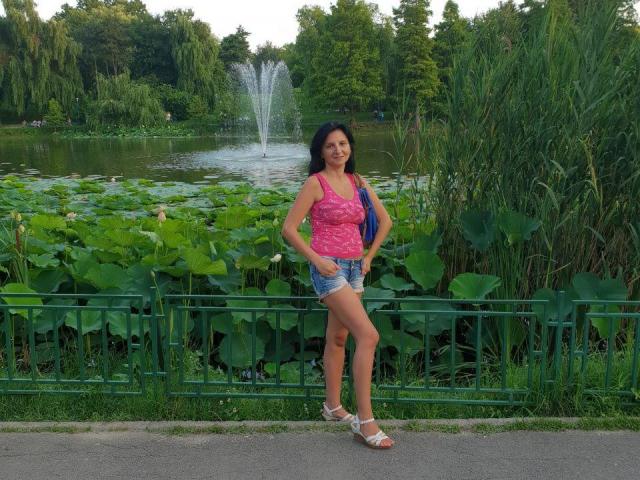 MadameAlexaX staat in een park, gekleed in een roze top, blauwe shorts en witte sandalen, met een waterfontein en groene planten op de achtergrond, met haar handen op haar heupen..