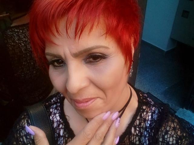 LadyLibely with red pixie hair, black lace top, and pink nails, looking slightly annoyed, holding her chin with her hand. Her tits are small, and her nails are long.