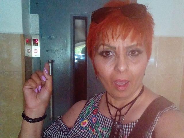 LadyLibely, met kort oranje haar, draagt zwarte zonnebril op haar hoofd, houdt een zilveren sigarettenhouder vast, draagt een zwart-wit geruit top met kleurrijke borduursels en een zwarte choker, en heeft volle lippen..