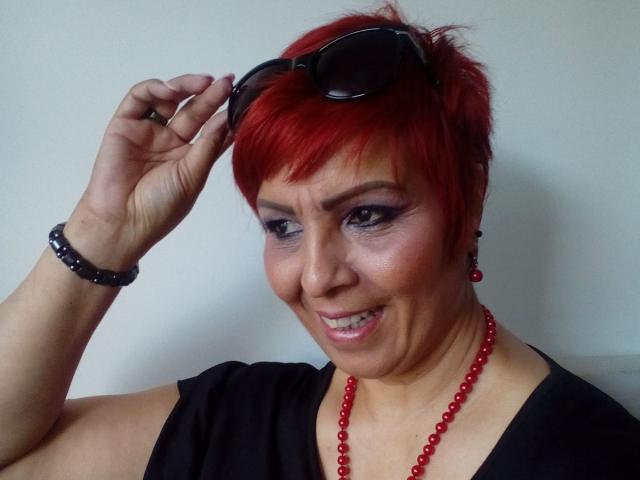 LadyLibely with short red hair, black sleeveless top, red bead necklace, black bracelet, holding sunglasses on head, smiling with gray eyeshadow and red earrings.