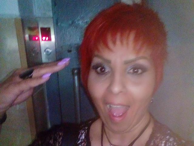 LadyLibely with short red hair, making a goofy face, tongue out, wearing dark makeup, and a black and white patterned top, hand with purple nails on the left.