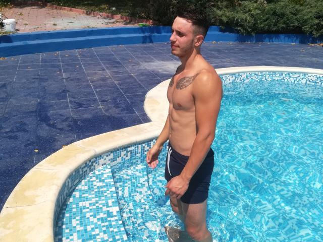 Steven Cum, senza camicia, è in una vasca idromassaggio con piastrelle blu, indossa pantaloncini da bagno neri, ha un tatuaggio sul petto destro e capelli corti e scuri. L'acqua schizza sulle sue gambe..