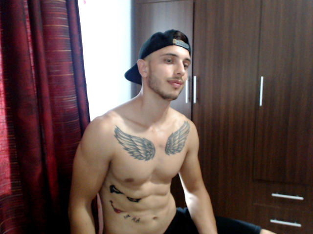 Shirtless StevenCum with black cap, wing tattoos on chest, and tattoos on left side, standing in room, brown wooden cabinets in background, red curtains on left.