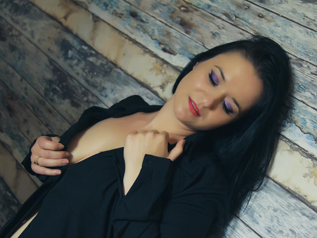 SensualCrissa, com cabelo preto, sombra roxa e batom vermelho, apoiada em uma parede de madeira, com uma camisa preta parcialmente aberta, revelando seus pequenos seios, e segurando a camisa com a mão direita..