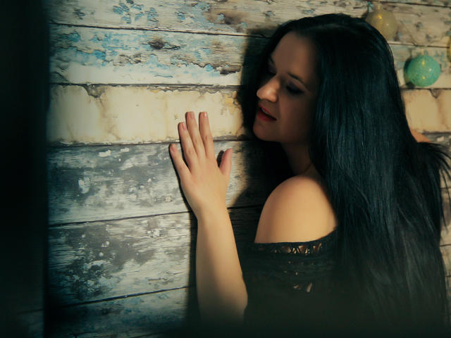 SensualCrissa with long black hair, red lipstick, and bare shoulders, touching a weathered wooden wall with her left hand, wearing an off-shoulder black top.