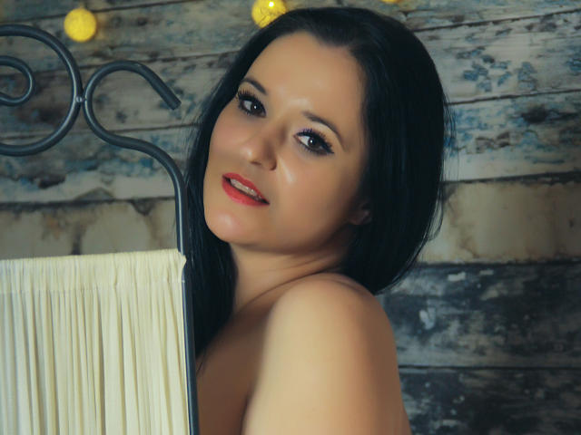 SensualCrissa with black hair, red lipstick, and dark eyeliner, looking up with a slight smile, her bare shoulders visible, standing in front of a metal frame with white cloth.