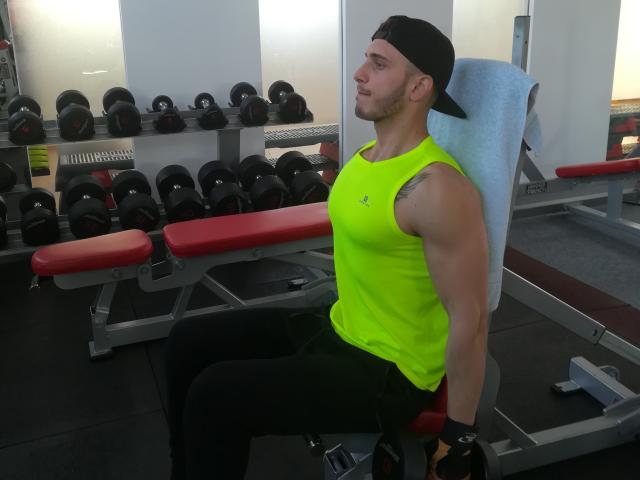 StevenCum, muscular with a tan, sits on a red bench in a gym, wearing a neon green sleeveless shirt, black pants, and a black backward cap, arms resting on bench.
