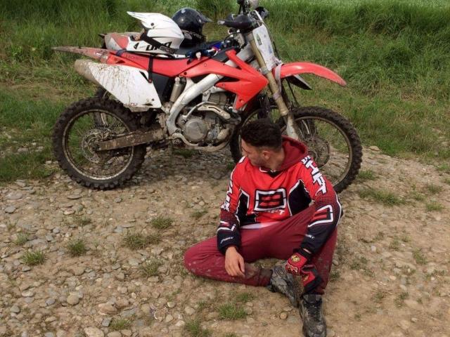 StevenCum kneels in front of a dirty red and white motorcycle, wearing a red and black motorcycle jersey and red pants, with a helmet on the bike.