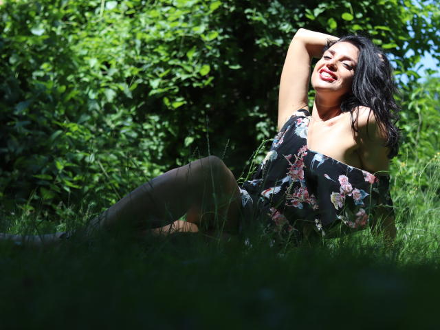 KatieFrenchie is a sexy chick with dark hair, red lipstick, and a floral off-shoulder dress, lying on grass, smiling, and showing her tits while raising an arm.