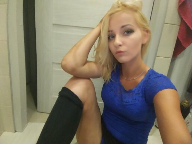 NatalieKiss with blonde hair, blue lace top, black knee socks, and a serious expression, sitting with one knee up, in a bathroom. Her tits are small, and she's cute.