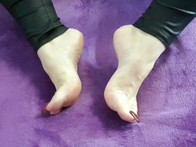 KaryQueen's bare feet with slightly curled toes, showing red nail polish, on a purple textured surface. She's wearing black pants, legs bent at the knees.