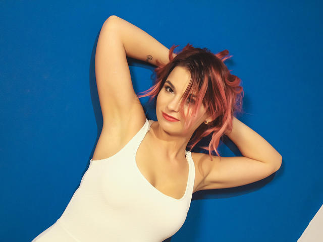 AliciaKristal, con cabello rizado rosa, camiseta blanca, brazos levantados mostrando las axilas, sonriendo, con un tatuaje en el antebrazo izquierdo, sobre un fondo azul, tiene senos pequeños, cara linda..