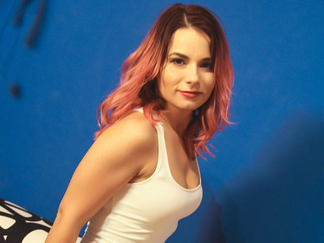 AliciaKristal, avec ses cheveux roses ondulés, portant un débardeur blanc, montrant ses seins, souriant à la caméra, sur un fond bleu..