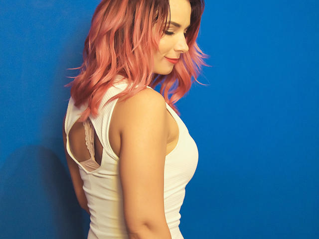 AliciaKristal con i capelli rosa ondulati, canotta bianca che lascia intravedere un reggiseno in pizzo, profilo laterale, sfondo blu, che guarda in basso con un leggero sorriso, mostrando il seno e un viso carino..