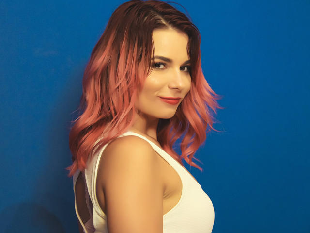 AliciaKristal, con i capelli rosa sfumati, una canottiera bianca e un sorriso adorabile, mostra leggermente il seno su uno sfondo blu. Sta guardando oltre la spalla, in modo sexy e sicuro di sé..