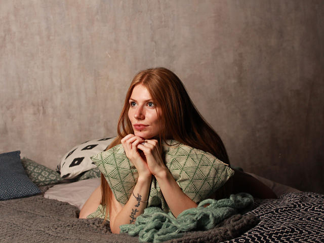 GretyArdent, avec de longs cheveux roux, tenant un oreiller vert à motifs, regardant l'appareil photo avec un léger sourire, allongée sur un lit avec des couvertures grises et vertes..