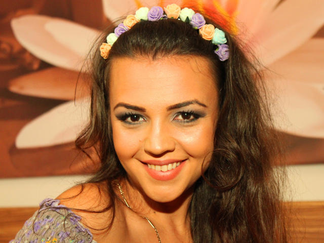 FunBrunettee with a flower crown in her brown hair, smiling at the camera. She has large tits, cute nose, and is wearing a purple dress. Bright, warm lighting.