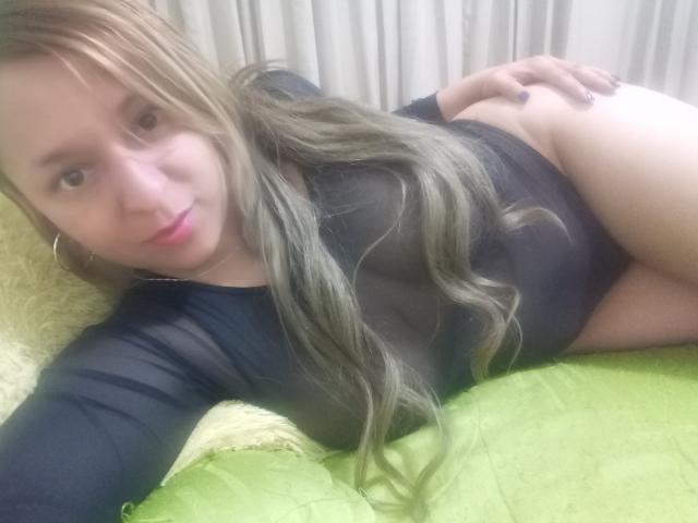Angelonix with long blonde hair, black top, lying on green bed, hand on butt, looking at camera, pink lips, hoop earrings, smiling, visible ass, soft lighting.