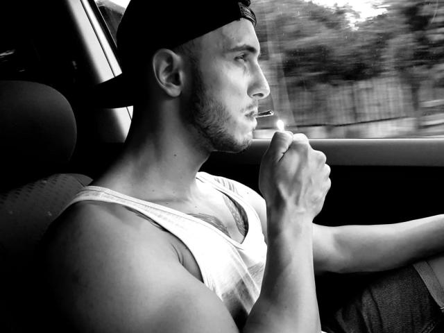 Black and white photo of StevenCum, muscular, smoking, wearing a backward baseball cap and white tank top, driving with right hand on wheel. Visible chest tattoo.