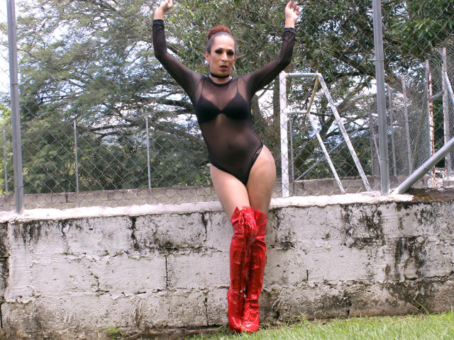 KhazandraX with red boots, black sheer long-sleeve bodysuit, and upraised arms, standing against a concrete wall, her tits visible through the mesh, her pussy covered.
