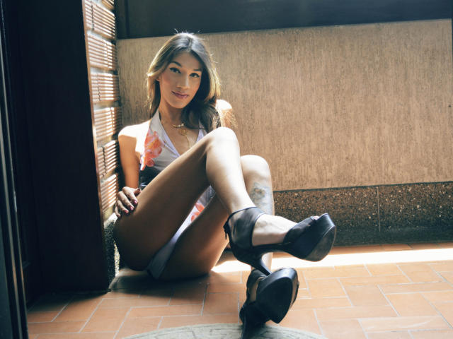Una joven morena con cabello castaño largo, vestida con un top blanco de tirantes, mostrando su trasero, y tacones altos negros, sentada en un piso de baldosas, con las piernas cruzadas, sonriendo, con un tatuaje en el muslo derecho..