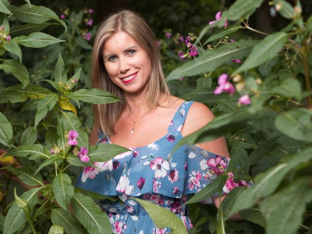 DoorenHot with blonde hair, blue floral dress, smiling, pink flowers, green leaves, and a cute necklace, standing in a lush garden. Her tits are partially visible.
