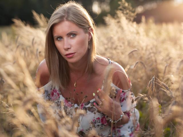 DoorenHot has blonde hair, blue eyes, and wears a floral off-shoulder top in a sunlit wheat field, holding a stalk, looking directly at the camera with a serious expression.