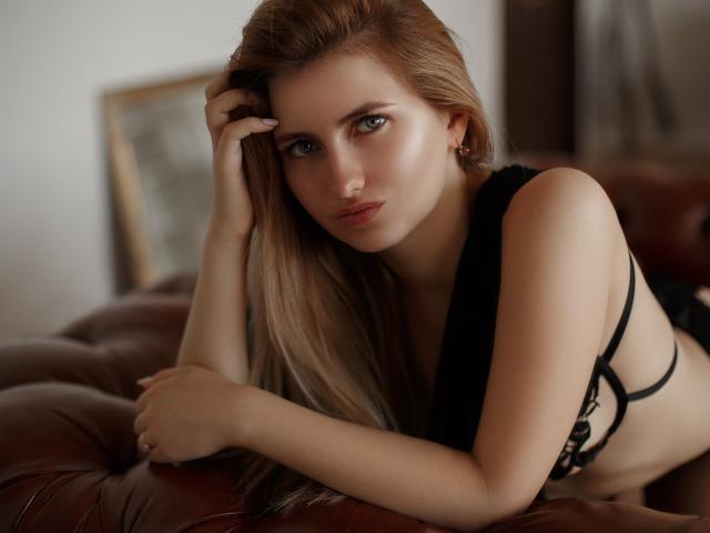 ShyNaomi with long blonde hair, black lace bra, and earrings, leaning on a brown leather couch, looking directly at the camera with a serious expression. Her tits are visible.