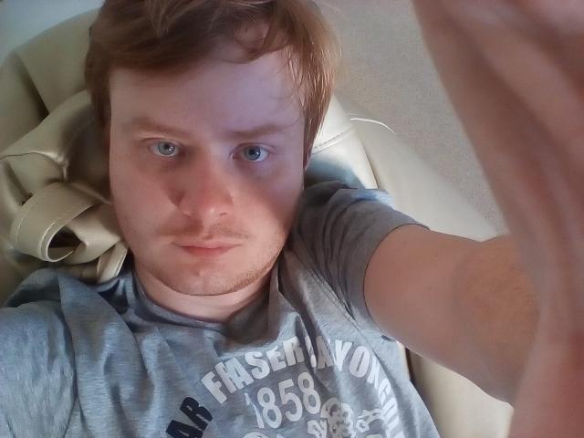 JeepersStrong tem pele clara, cabelo ruivo, olhos azuis e uma barba clara. Ele está vestindo uma camisa cinza, deitado em uma cadeira de couro bege, com o braço direito dentro do quadro..