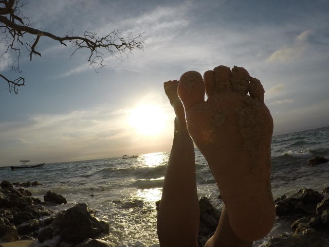 Os pés arenosos de PiedsMagiques em primeiro plano, pernas levantadas, sol ao fundo, ondas do oceano, rochas e um galho de árvore no canto superior esquerdo. A luz do sol reflete na água..