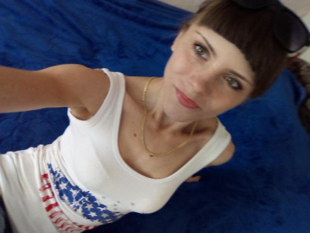 MilaDelight, con pechos pequeños, vistiendo una camiseta blanca con un diseño de la bandera estadounidense, un collar dorado y flequillo, tomando una selfie desde arriba. Fondo de tela azul..