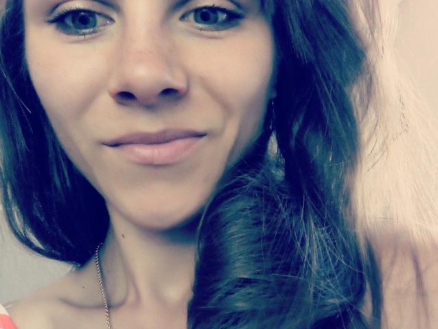 MilaDelight has light skin, blue eyes, and brown wavy hair, with a subtle smile and no makeup, showing her cute nose and small tits. She's wearing a silver chain.