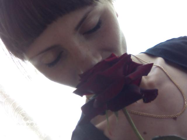 MilaDelight with dark brown bangs, eyes closed, smelling a red rose, wearing a gold necklace, black top, and pale skin. Her nose is near the rose's petals.
