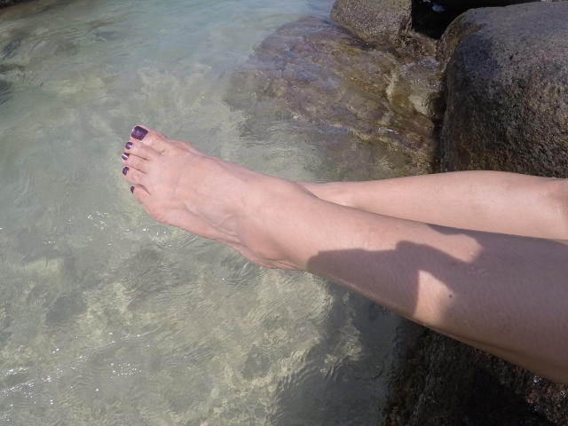 PiedsMagiques', com suas pernas pálidas e esguias, com os dedos pintados de roxo em água clara e rasa, as pernas apoiadas em uma superfície rochosa, com a luz do sol projetando uma sombra..