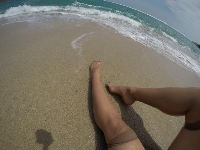 Les jambes bronzées de PiedsMagiques s'étendent sur la plage de sable, les orteils dans une douce vague, les cuisses légèrement visibles, une ombre sur le sable, l'océan en arrière-plan, les pieds pointés vers l'avant, une journée calme..