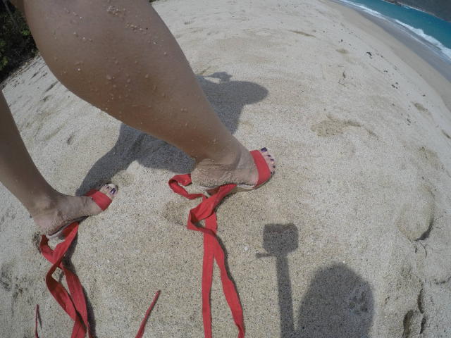 As pernas cobertas de areia de PiedsMagiques, usando chinelos vermelhos com fitas, estão em pé em uma praia arenosa, mostrando sua sombra e uma pequena sombra de uma câmera na areia..