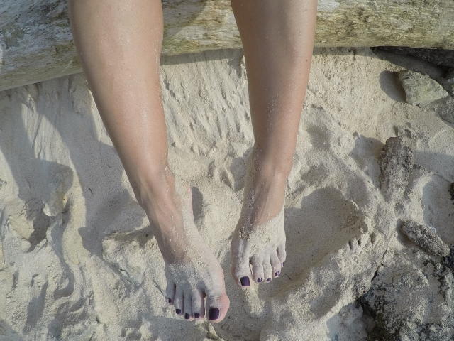 De benen en voeten van PiedsMagiques zijn bedekt met zand, met paars geschilderde tenen, en staan op een rotsachtige, zandige ondergrond. Haar huid is bleek, en haar voeten staan iets uit elkaar..