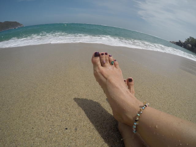De voeten van PiedsMagiques, met paarse nagellak, een groene ring om de grote teen en een kralen enkelbandje, rusten op een zandstrand, met een blauwgroene oceaan op de achtergrond en een heldere lucht erboven..