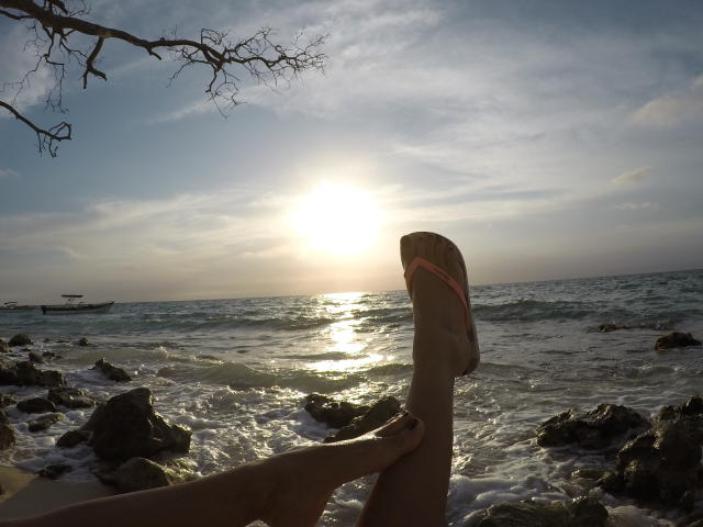 Le gambe di PiedsMagiques sono in posizione eretta, con un piede infilato in un ciabatta arancione, su una spiaggia rocciosa, con il sole che splende, le onde dell'oceano e un ramo senza foglie nell'angolo in alto a sinistra..