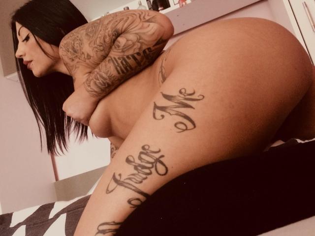 SweetAnnalBelle desnuda, con tatuajes en los brazos y el muslo, inclinada hacia adelante con el trasero en el aire, cabello negro, perfil lateral, mirando hacia abajo, iluminación suave, ambiente de dormitorio..