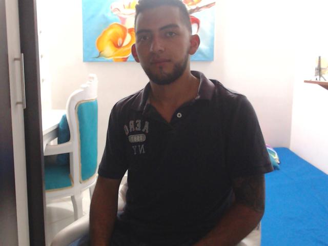 SteveBigCock, con cabello negro corto, barba, tatuaje en el brazo derecho, vistiendo una camisa polo oscura, de pie en una habitación con alfombra azul, una silla blanca y arte de pared colorido..