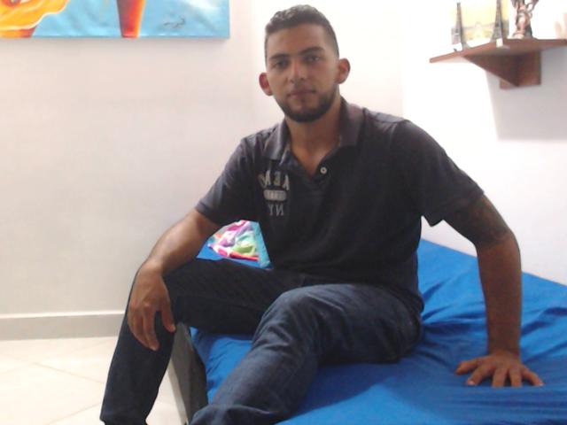 SteveBigCock, zittend op een blauw bed, gekleed in een donker poloshirt en jeans, heeft een getrimde baard en kort haar, met een tatoeage op zijn rechterarm..