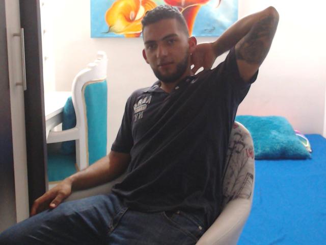 SteveBigCock, avec des tatouages sur le bras, portant une chemise noire et un jean bleu, avec une courte barbe, assis sur une chaise, le bras droit levé, regardant la caméra, lit en bleu, chaise blanche, décoration murale colorée..